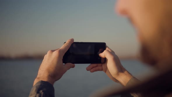 Man Hands Holding Mobile Phone And Taking Picture At Sunset. Guy Taking Photo of Sunset With Mobile. alt