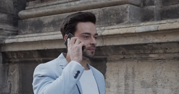 Young Man Rides Near The Ancient Castle In A Festive Light Suit And Talks On The Phone alt