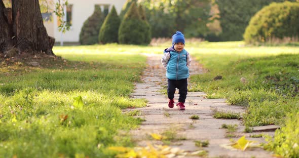 Alone Toddler in Blue Hat and Vest Walking Away Funnily in Autumn Park