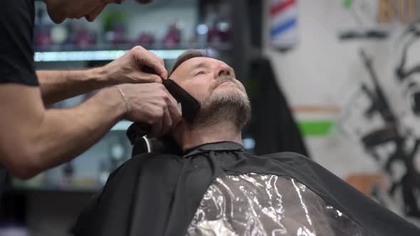 Barber master shaving handsome mature bearded man using electric shaver in salon.