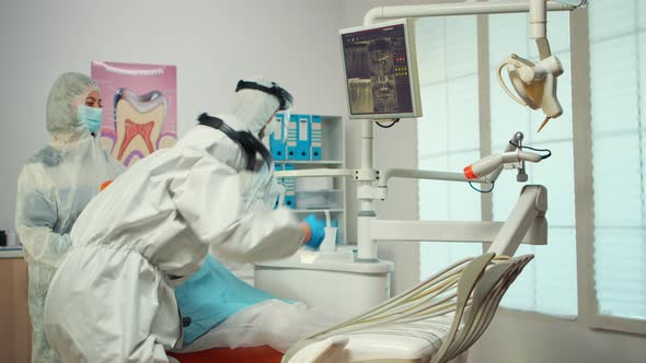 Dentist in Ppe Suit Pointing on Digital Dental Monitor Explaining Xray To Mother alt