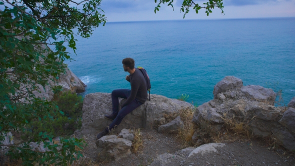 Young Hipster Guy Sitting On The Rock Mountains Near Sea, Man With a Rucksack On His Back alt
