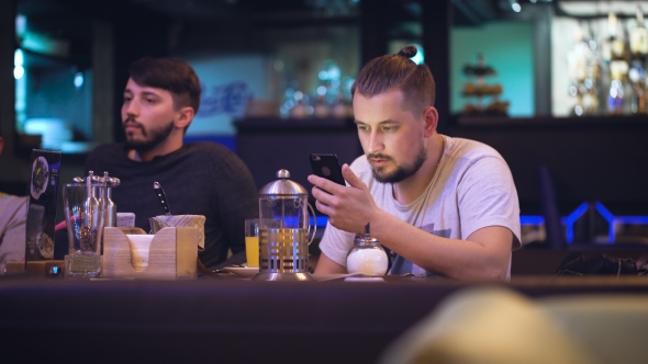 Cute Hipster Sits At a Table In a Nightclub, And Prints Something On The Smartphone. alt