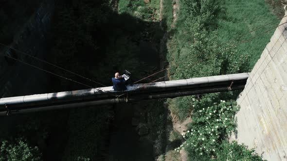 Businessman sitting on pipeline over trench using laptop and phone alt