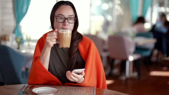 Brunette Woman is Drinking Coffee Restaurant Viewing Video or Pictures in Smartphone alt