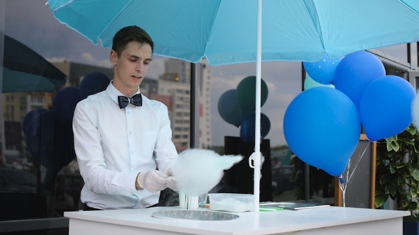 Young Guy Making Cotton Candy On a Special Machine, It Wears Bow Tie, Behind Him Balloons