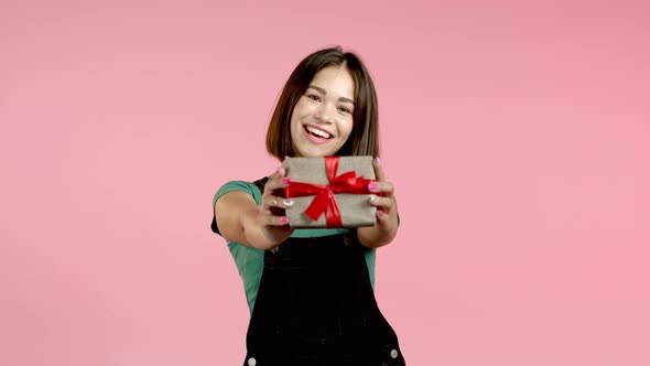 Excited Woman Holding Gift Box and Gives It By Hands To Camera on Pink Wall Background. Girl Smiling alt