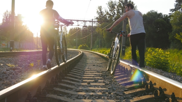 Two Male Friends Walking With Their Bikes On The Rails At The Railway In Sunset And Talking.