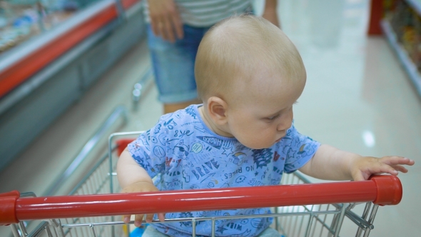 Little Baby Sitting in a Grocery Cart in a Supermarket, While His Father Chooses Purchases. alt