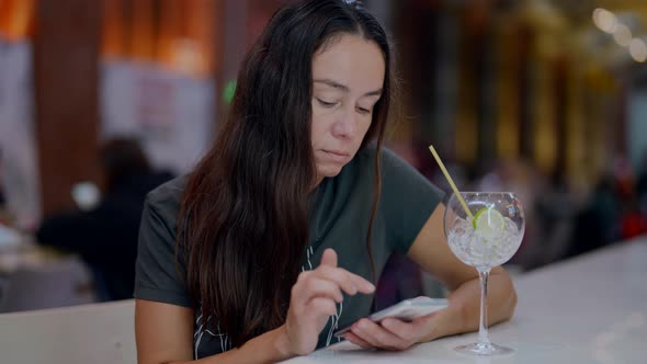 a Brunette with Long Hair is Sitting at the Counter with a Glass and Checking Phone alt