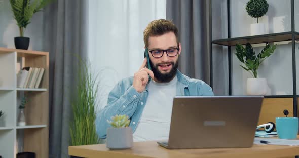 Male Worker in Glasses which Sitting in Front of Computer at Home and Talking on mobile alt