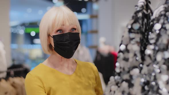 Mature Woman Shopper in Protective Mask in Clothing Store Admires Shiny Outfit Elderly Lady Looks alt