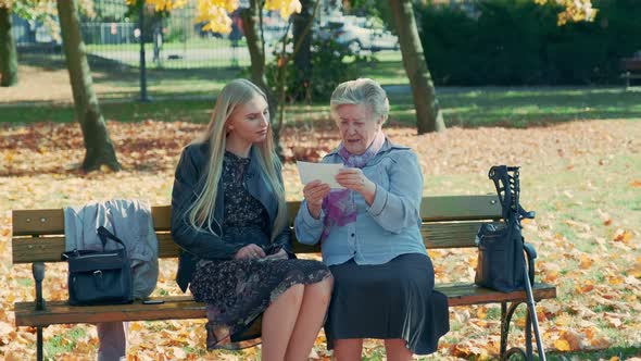 Young Blonde Woman Listening To and Old Woman Reading Something While Sitting Together on a Bench alt
