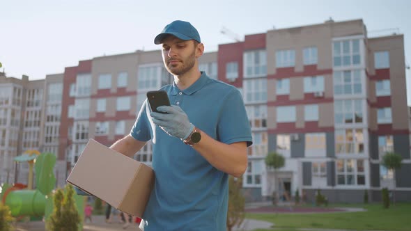 The Postman with Glasses Carries the Parcel and Looks at the Delivery Address Via Mobile Phone alt
