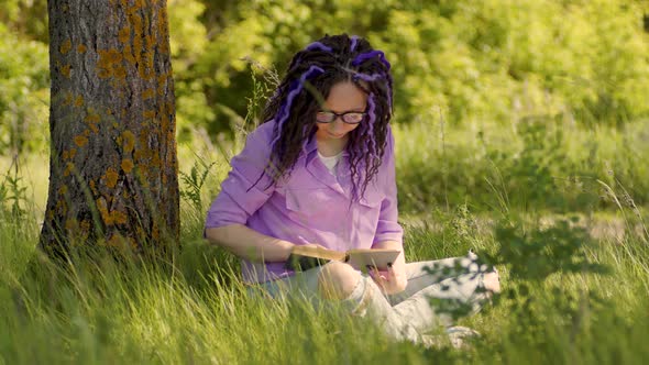 A woman in a purple shirt and glasses reads a literature under a tree. alt