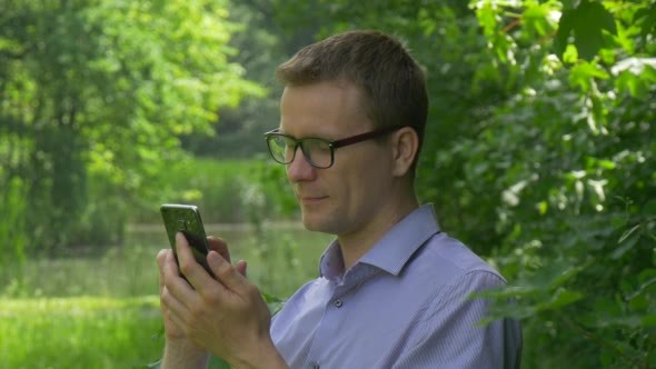 Man Clicks Mobile Phone in Park Smiling Texting