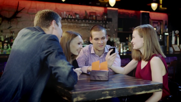 Company Of Young People Is Watching Something In The Cellphone Sitting In The Bar