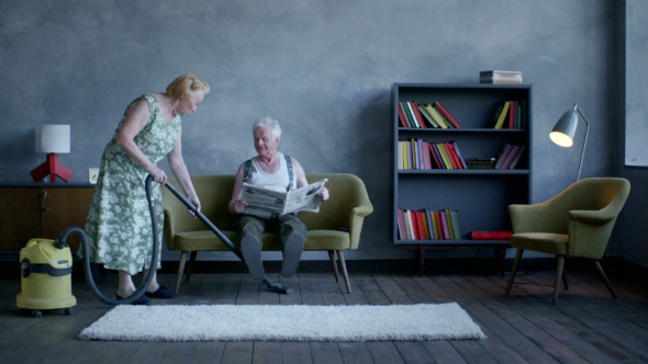Elderly Woman Vacuuming The Floor, And An Elderly Man Reading a Newspaper alt