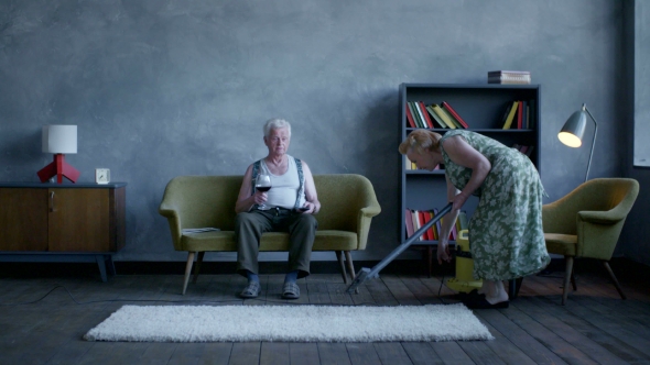 Senior Couple Quarrel. Husband Drinking Wine, His Wife Cleans The House And Scolds Him alt