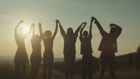 Silhouette of Diverse Multiracial Friends Standing with Arms Raised on Mountain Peak at Sunset alt