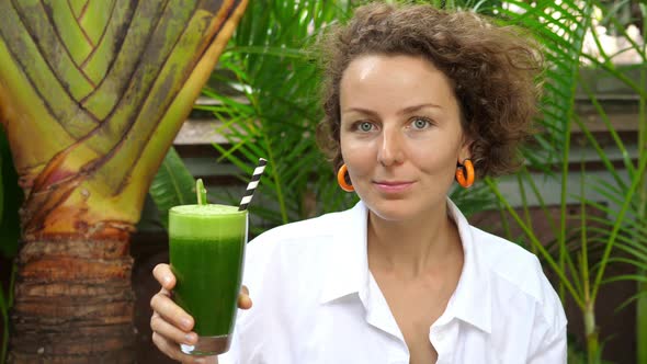 A Young Caucasian Woman Holds a Organic Cold Pressed Green Juice and Looking to the Camera alt