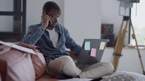 Busy Young African American Man Receiving Phone Call Sitting on Couch in Home Office with Laptop alt