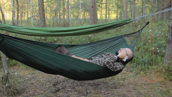 A Man In a Hammock Is Calling On The Phone. Man In The Woods With a Beard And Glasses