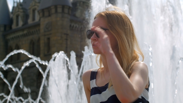 Smiling Blonde Is Sitting On a Fountain Near a Castle And Touching Sunglasses