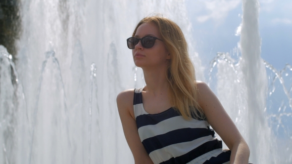 a Smiling Blonde In Sunglasses Is Sitting On a Fountain, Looking At Camera
