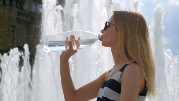 Blonde in Sunglasses is Drinking Water From Plastic Bottle Sitting on a Fountain