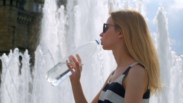 Blonde Dressed In a Striped Dress And Sunglasses Is Drinking Water From Bottle