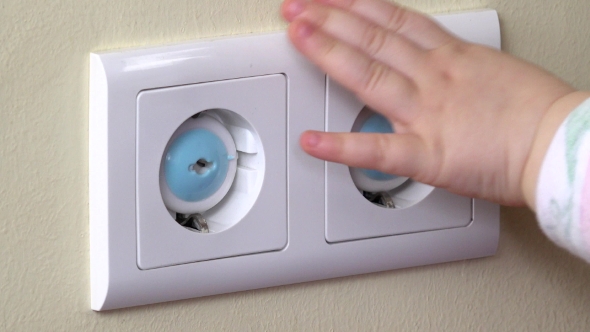 Baby Hand Reaching For An Electrical Outlet Covered With Blue Safety Plugs