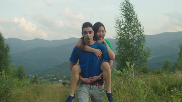 Carefree Arab Man Piggybacking Joyful Excited Asian Girlfriend in Mountains alt