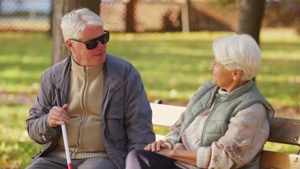 Blind Elderly Man Sitting with His Wife on the Bench in the Park and Holding Blind Cane alt