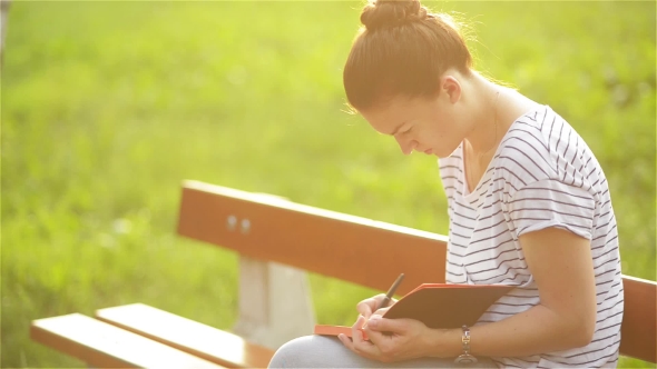 Beautiful Young Woman Is Writing a Diary Outdoors In The Park, Student Studying On a Bench alt