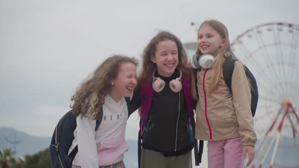 Three School Girls with Backpacks Laugh Hard with Ferris Wheel on Background