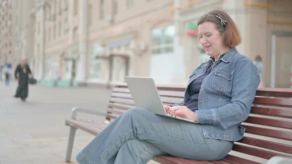 Old Woman Talking on Video Call While Sitting Outdoor on Bench alt