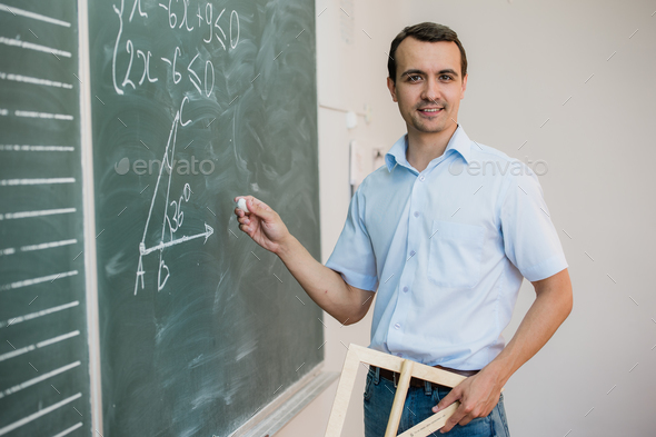 Young teacher or student holding triangle pointing at chalkboard with formula, looking to camera and