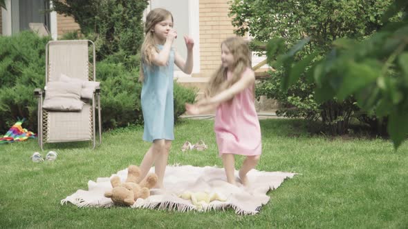 Two Cheerful Little Girls Taking Each Other's Hands and Start Jumping on Blanket Outdoors. Wide Shot alt