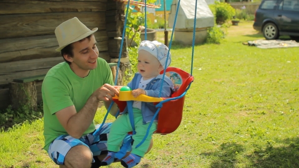 Father Rolls The Beautiful Baby On a Swing. Yang Man With His Son In The Garden Of His Home alt