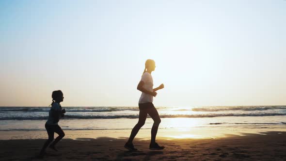 Mom and Daughter are Jogging on the Sea Sand Beach at Sunset alt