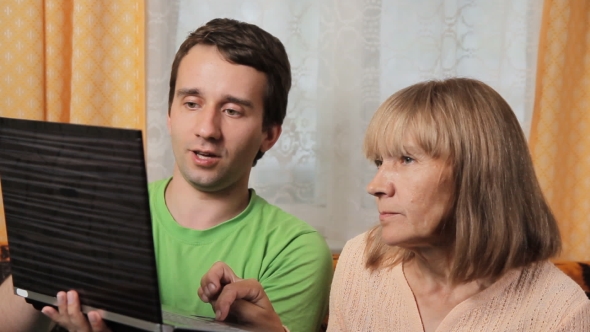 Adult Male Teaches Old Woman Working For a Laptop. Mother And Son Sitting On a Sofa At Home