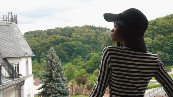 Sexy African American Lady In Black Hat And Fashionable Striped Dress Standing On The Balcony