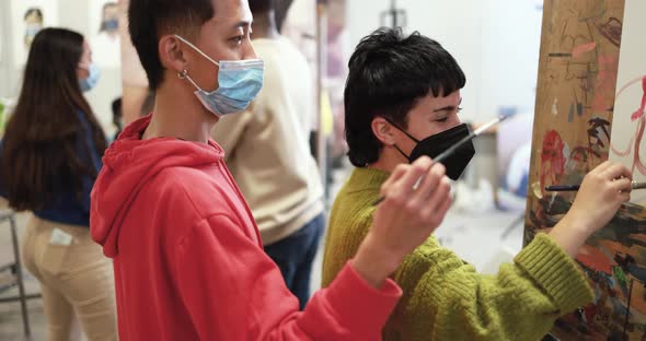 Multiracial students painting inside art room class wearing safety masks alt