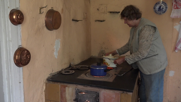Elderly Woman Put Rustic Pot On Rural Clay Stove In Kitchen.