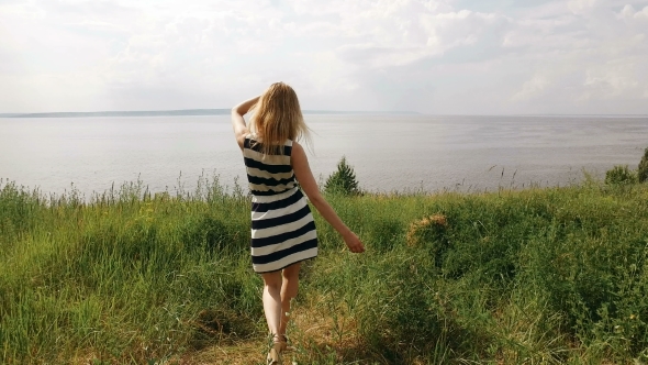 Young Shapely Blonde Is Walking On a Field, Looking On a Sea In Summertime