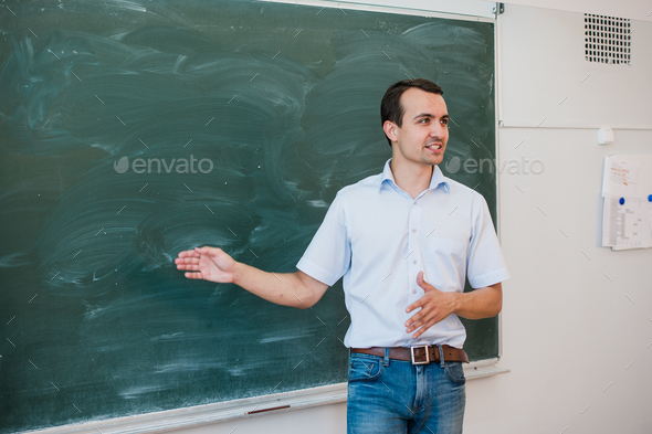 Portrait of young handsome student or teacher in a class pointing at blank chalkboard, talking and