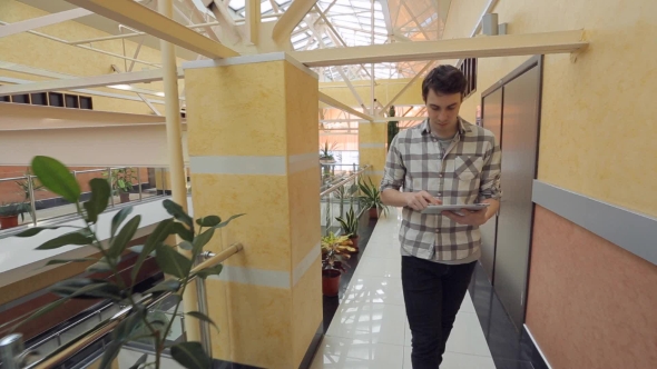 Young Man Walks Through Shopping Mall With Tablet In His Hands. alt