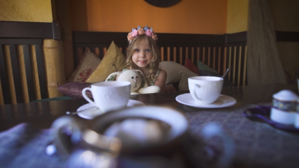 A Little Happy Girl Is Sitting On The Couch At a Cafe And Hugging Her Stuffed Rabbit. alt