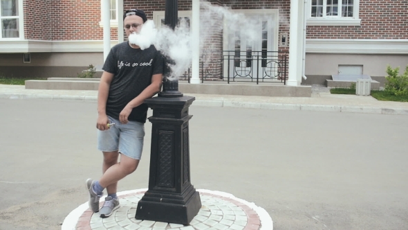 Young Man Standing At Street Lamp And Smoke E-Cigarette. Vape. 